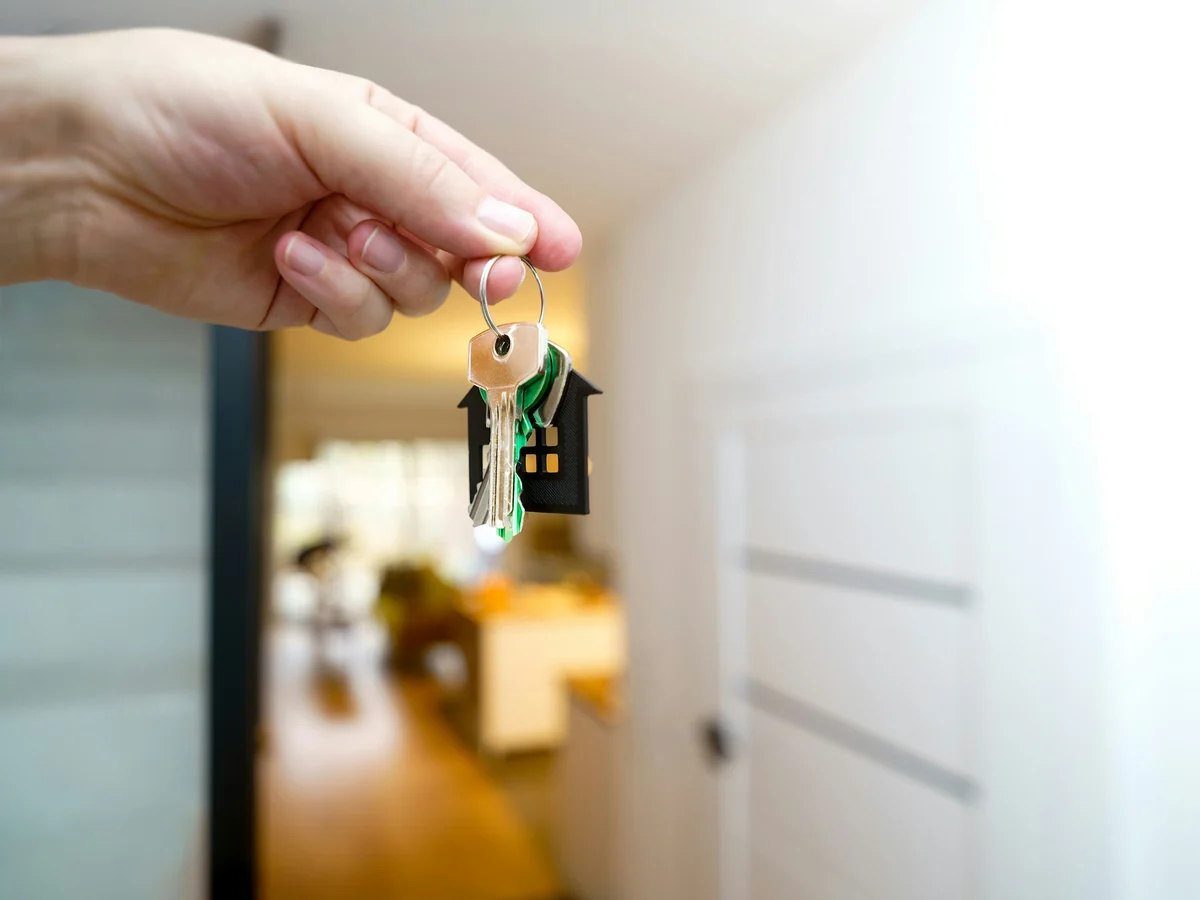 Hand holding a set of house keys with a small house keychain inside a bright home
